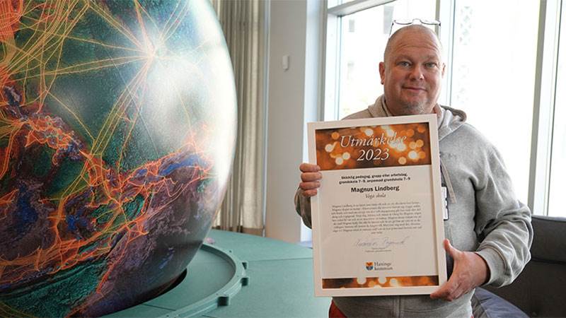 Magnus Lindberg, pedagog vid Vega skola. Man håller i diplom ståendes framför en jordglobsskulptur inne i biblioteket på Vega skola.