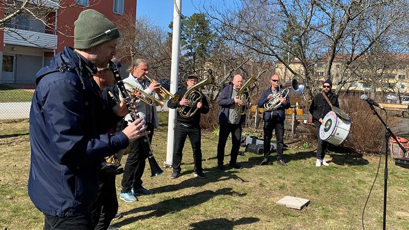 Süperstar Orkestar gästar Vallagården i Handen. Åtta män står i halvcirkel på gräsmattan på Vallagården och spelar instrument.