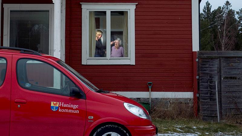 En hemtjänstbil står parkerad utanför ett hus. I husets fönster syns en äldre och en yngre kvinna.