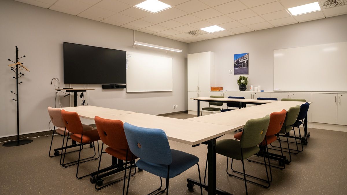 Ett mötesrum möblerat med bord i U‑form, omgivna av stolar i olika färger samt utrustat med skärm, whiteboard och förvaring.