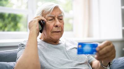 Äldre man sittandes med telefon och tittar på sitt kontokort.