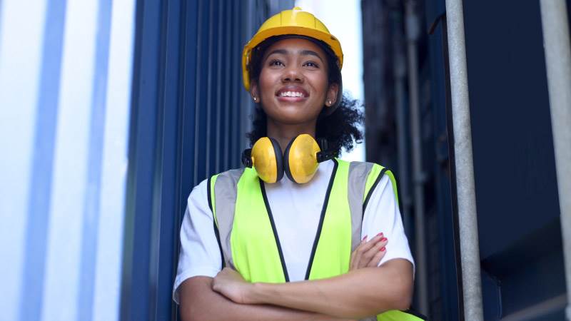 Ung kvinna ståendes på arbetsplats med bygghjälm på huvudet.