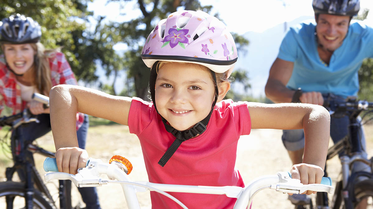 En glad flicka i cykelhjälm håller händerna på sitt cykelstyre i ett fast grepp och ser in i kameran. Bakom  henne kommer en glad kvinna och man i cykelhjälmar på varsin cykel.
