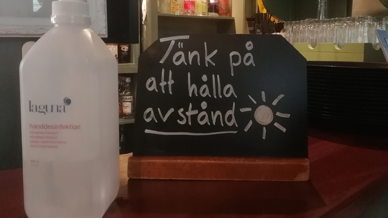 En flaska handdesinfektion och en skylt med  texten "Tänk på att hålla avstånd" står på en bardisk i en restaurang.