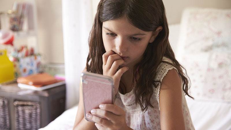 Flicka sitter på sängen i sitt rum och tittar på en mobiltelefon