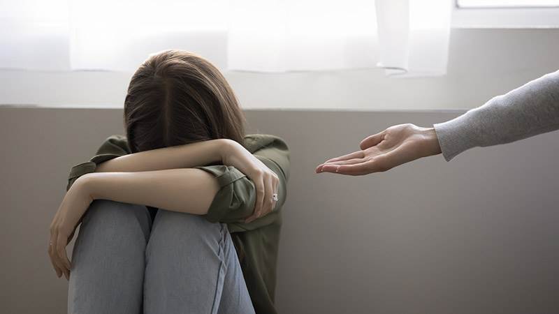 Ung tjej vilar huvudet i sina armar, sitter hopkrupen mot en vägg. En hjälpande kvinnohand sträcks ut mot den unga tjejen.