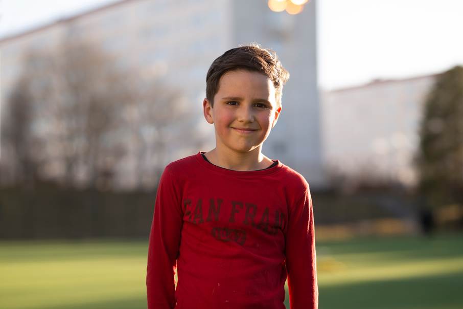 Ung pojke i röd tröja ser in i kameran där han befinner sig på en fotbollsplan i Brandbergen.