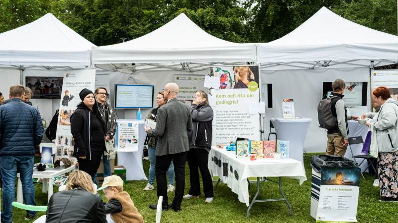 Haninge kommuns förskolor och skolor på Haningedagen 2022. Vita utställningstält på en grön gräsmatta. Vuxna står och samtalar utanför tältet. En liten pojke och mamma sitter vid ett bord och målar.