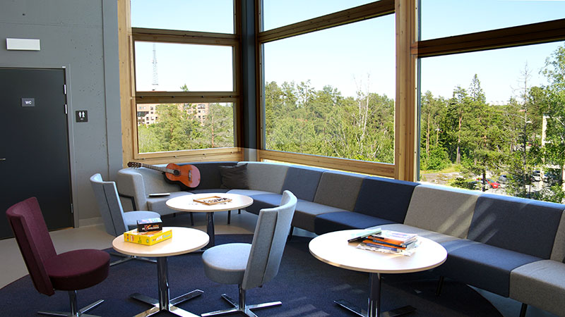Ett uppehållsrum med lång blå soffa små bord och stora fönster.
