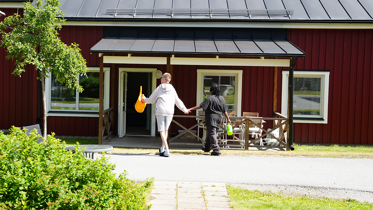 Två personer går mot ett hus med vattenkannor i handen.