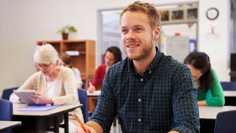 En man sitter i skolbänken i ett klassrum och antecknar