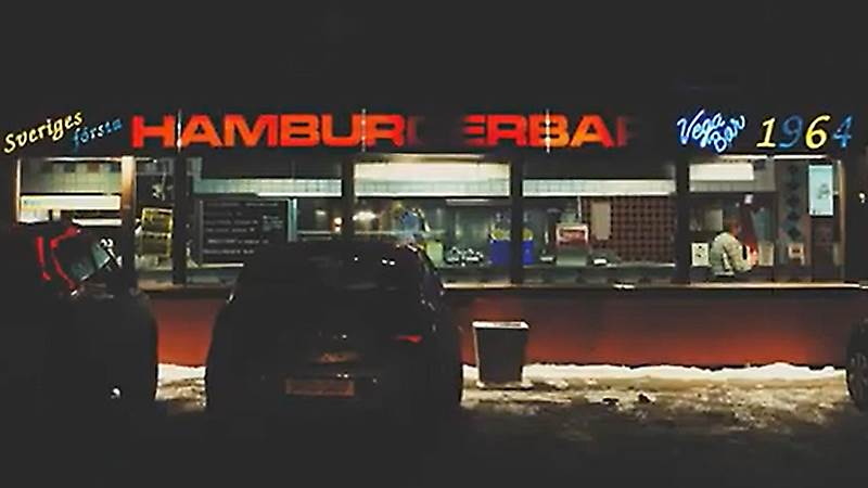 Vegabaren Framsidan på Vegabarens under en mörk kväll. Två bilar står parkerade utanför och ovanför ett stort fönsterparti står med upplysta bokstäver "Sveriges första hamburgerbar Vega bar 1964".