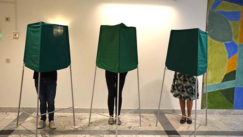 Tre personer ståendes bakom gröna skärmar i en vallokal.