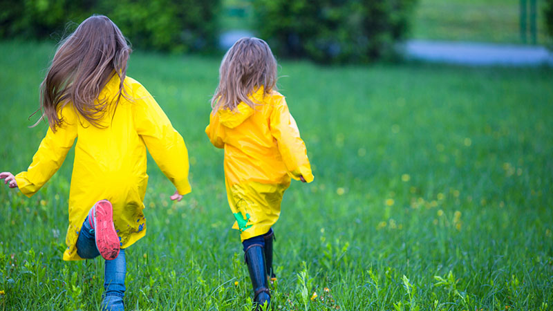 Två tjejer i gula regnrockar springer över en gräsmatta