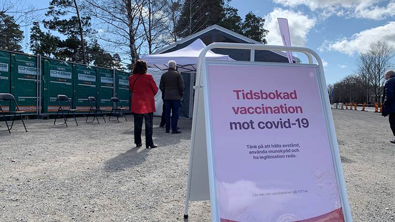 En kö utanför vaccinationsmottagningen vid Torvalla i Haninge. I förgrunden står en gatupratare med texten "Tidsbokad vaccination mot covid-19".