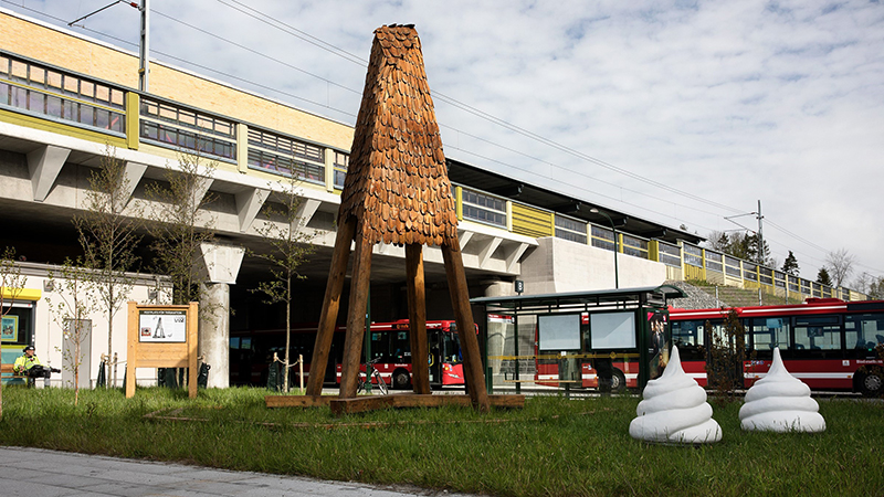 Maränger vid en hög träskulptur intill bussterminal.