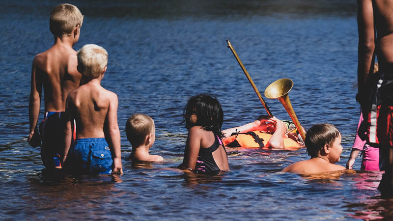 Blåsartrion FriLuft som spelar på förra årets Sommar på Rudan i en sjö, iklädd flytväst och med barn runtomkring.