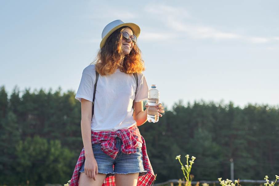 Tonårsflicka står med en flaska vatten i handen med solglasögon i solen. 