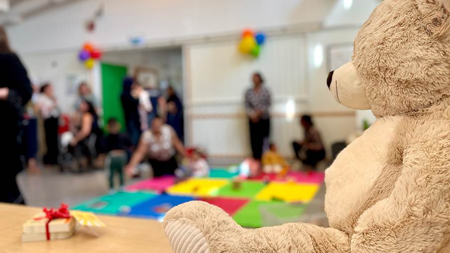 Familjecentralen Jordbro håller öppet på fredagar. Nallebjörn i förgrunden, i bakgrunden barn och vuxna.