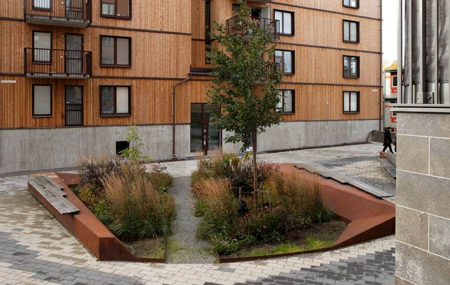 Grönskande dagvattenlösning. som ger plats för vila och återhämtning. Ingrid Horns torg, Handen. Arkitekt: Cedervall arkitekter Växtlighet vid ett litet torg omgärdat av gatustenar emd en mörk träfasad i bakgrunden