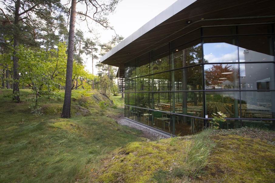 Kulturhusets läsesal. Till höger i bild glasfönster med läsplatser på insidan, till vänster mossbeklädda hällar med grönt gräs och träd omkring.dda d läsplatser på insidan, till vänster