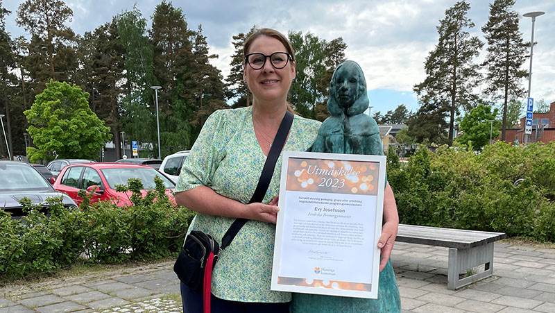 Evy Josefsson, lärare vid Fredrika Bremergymnasiet. Kvinna håller i diplom intill en staty utomhus. Foto i halvfigur.