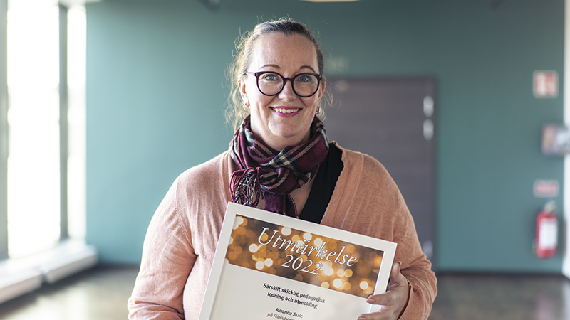 En glad kvinna i  glasögon och rosa tröja håller ett diplom