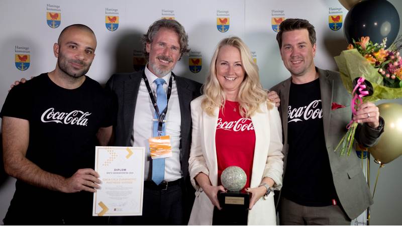 Coca-Cola Europacific Partners Sverige AB och Sven Gustavsson (M), kommunstyrelsens ordförande Tre representanter från Coca Cola fotade framför en fotovägg tillsammans med Sven Gustafsson. Diplom, ballonger och pris syns i bildr