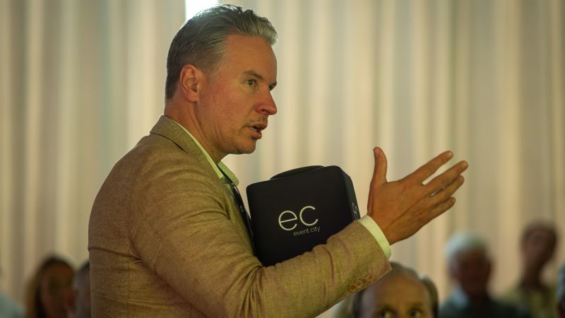 Man i kavaj  står upp bland åhörarna och talar