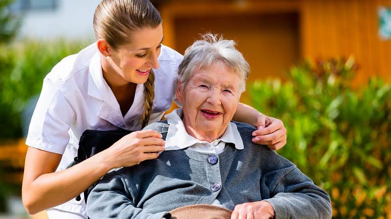 En glad äldre kvinna sitter i rullstol. En sköterska står bakom och lutar sig framför kvinnan i rullstol för att säga något.