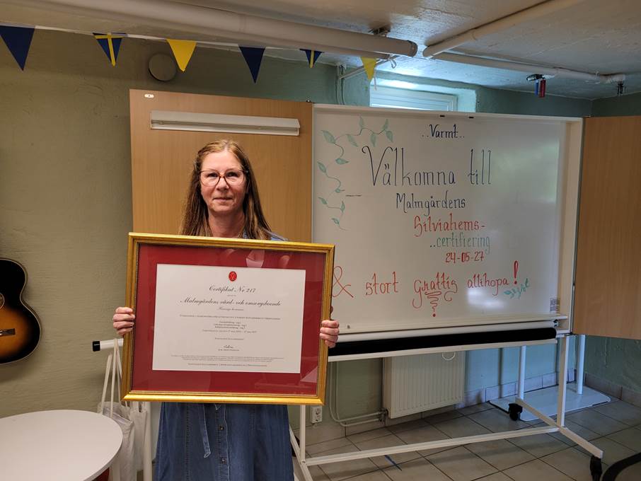 Malmgårdens verksamhetschef Maria Mattsson visar stolt upp boendets certifikat från Silviahemmet. Bild på en kvinna som håller i ett guldinramat diplom.