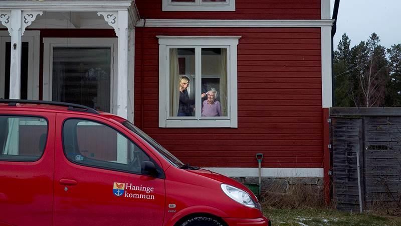 En röd hemtjänstbil med Haninge kommuns logga på står utanför ett rött hus.