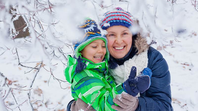 En vinterklädd, glad pojke och mamma under en snöig trädgren