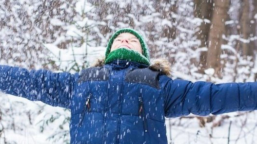 En ung person i mössa och vinterjacka stäcker ut armarna och njuter av snöflingor som seglar ner från skyn.