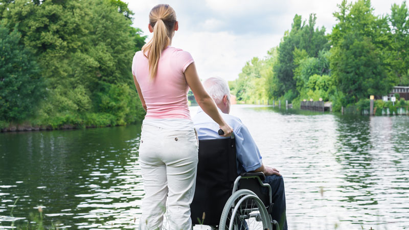 En man sitter i en rullstol och ser ut över en sjö. En kvinna håller i rullstolen.