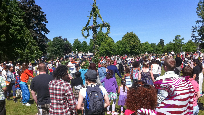 Människor dansar runt en midsommarstång klädd med gröna löv  i Jordbro
