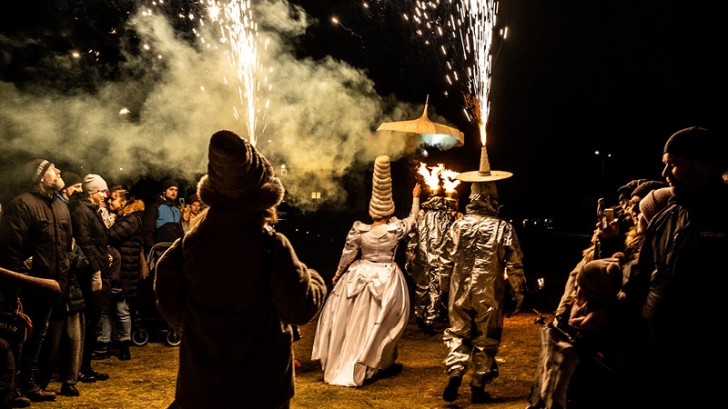 En kvinna i stor, puffig klänning och två män i silverdräkter ger en eldsghow framför publik.