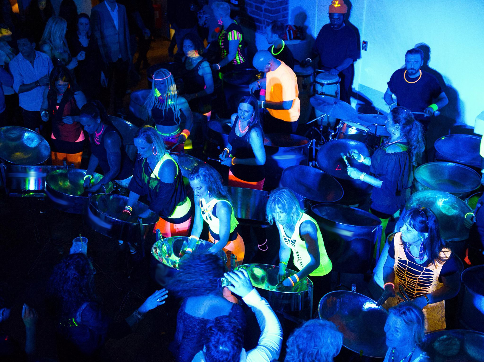 Stockholm Steelband Bild på medlemmarna i Stockholm Steelband i en blåfärgad lokal där kläderna lyser i neon.