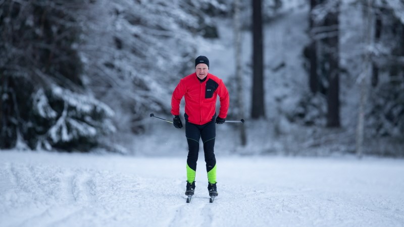 Äldre man i röd tröja åker skidor. Snöiga träd syns i bakgrunden.